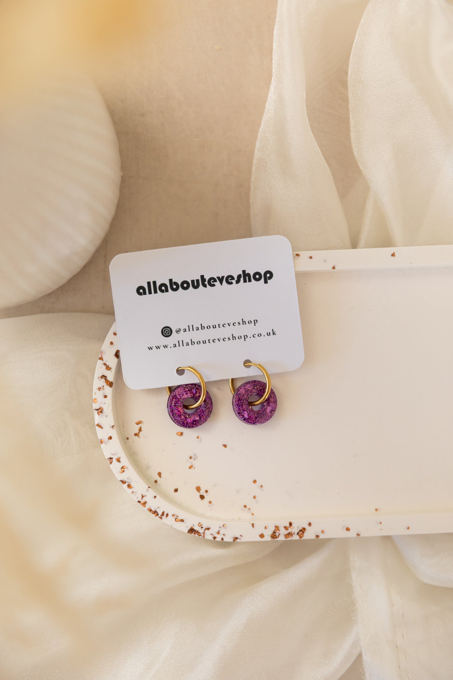 Pair of purple earrings on a white card with 'allabouteveshop' branding, displayed on beige fabric.