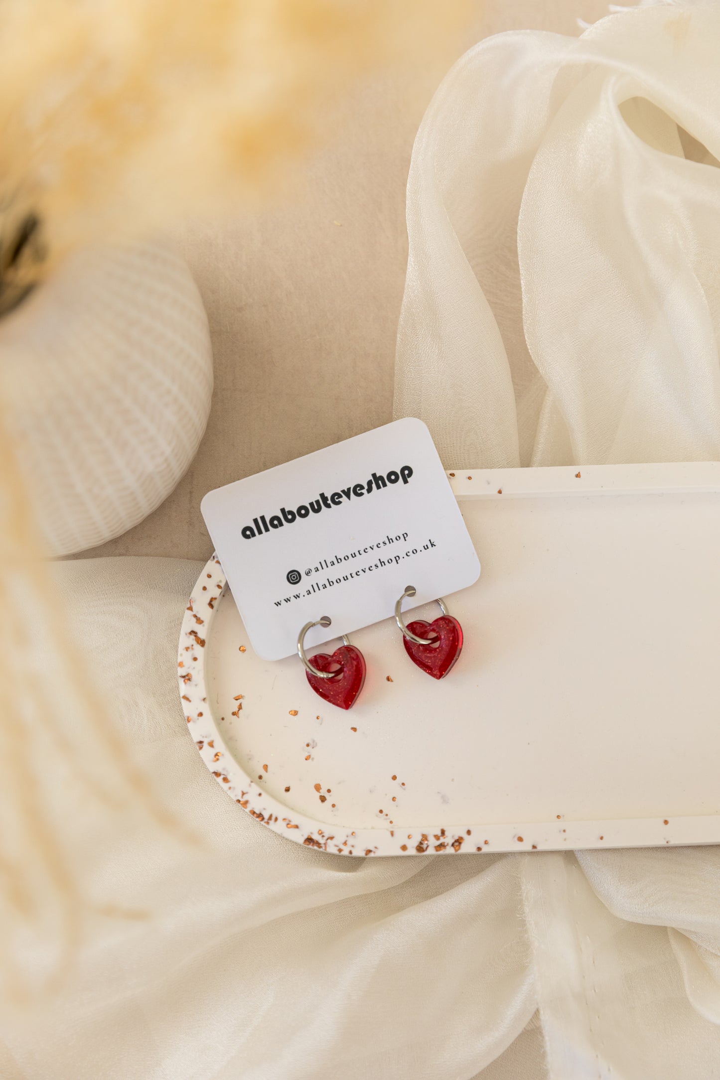 Heart-shaped earrings on a white card with 'allabouteve' branding, against a soft beige fabric background.