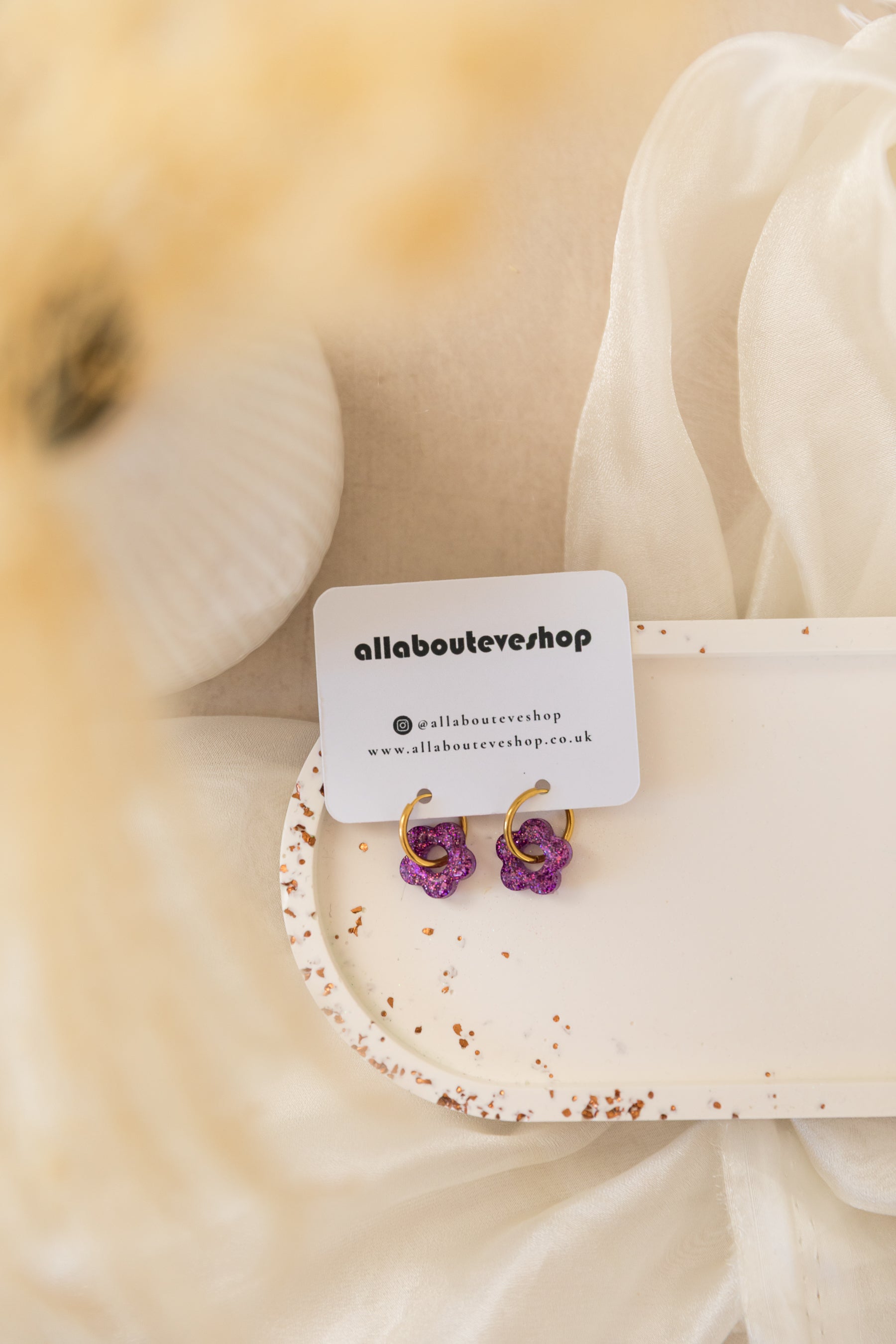 Pair of purple flower earrings on a white card with 'allaboulevardshop' branding, against a blurred floral background.