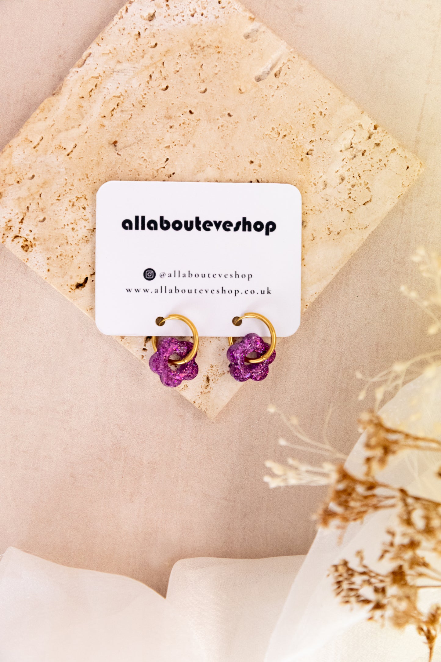 Purple earrings on a card with 'allabouteveshop' branding, placed on a textured beige surface.