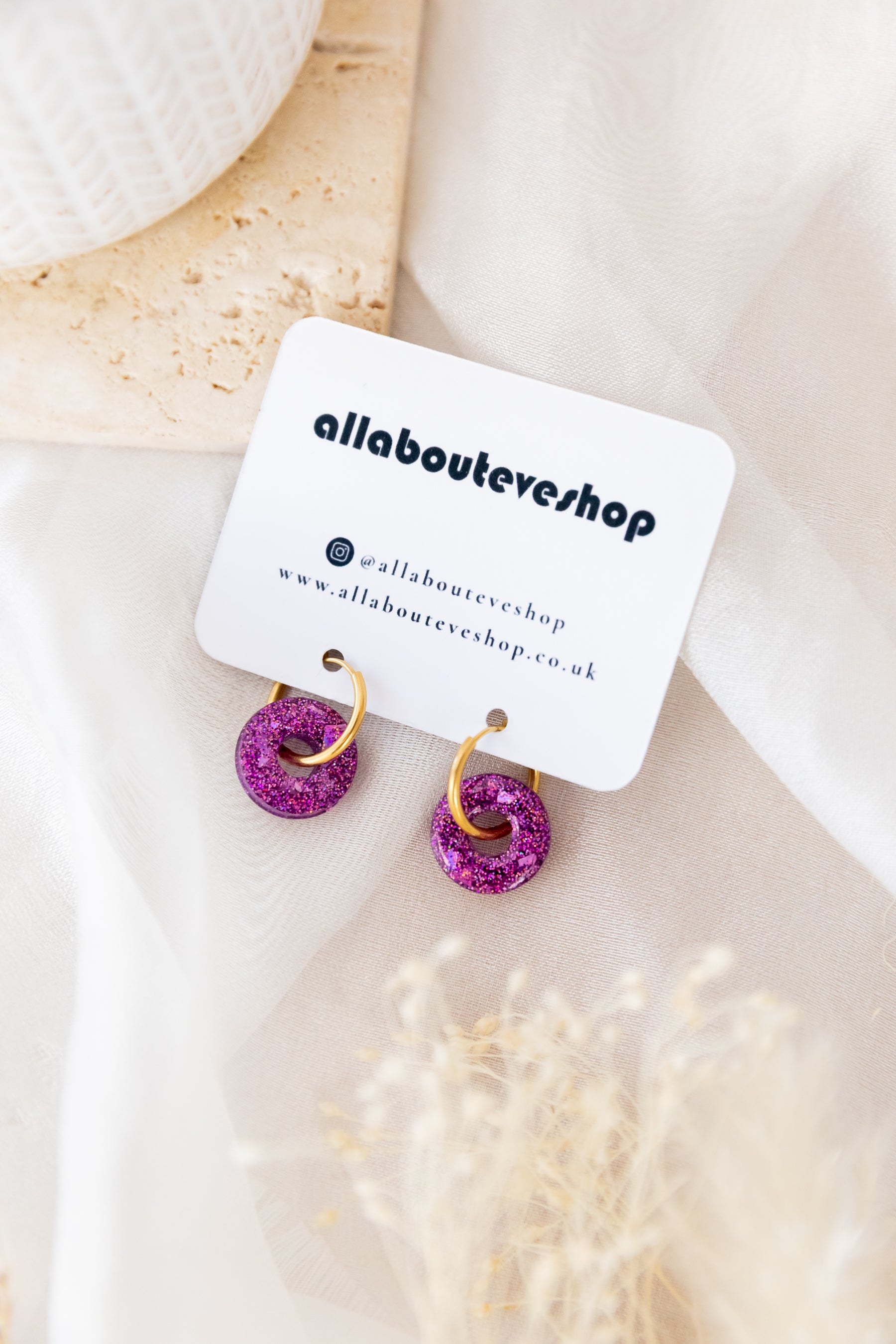 Pair of earrings with pink and gold details on a branded card, placed on a light surface.