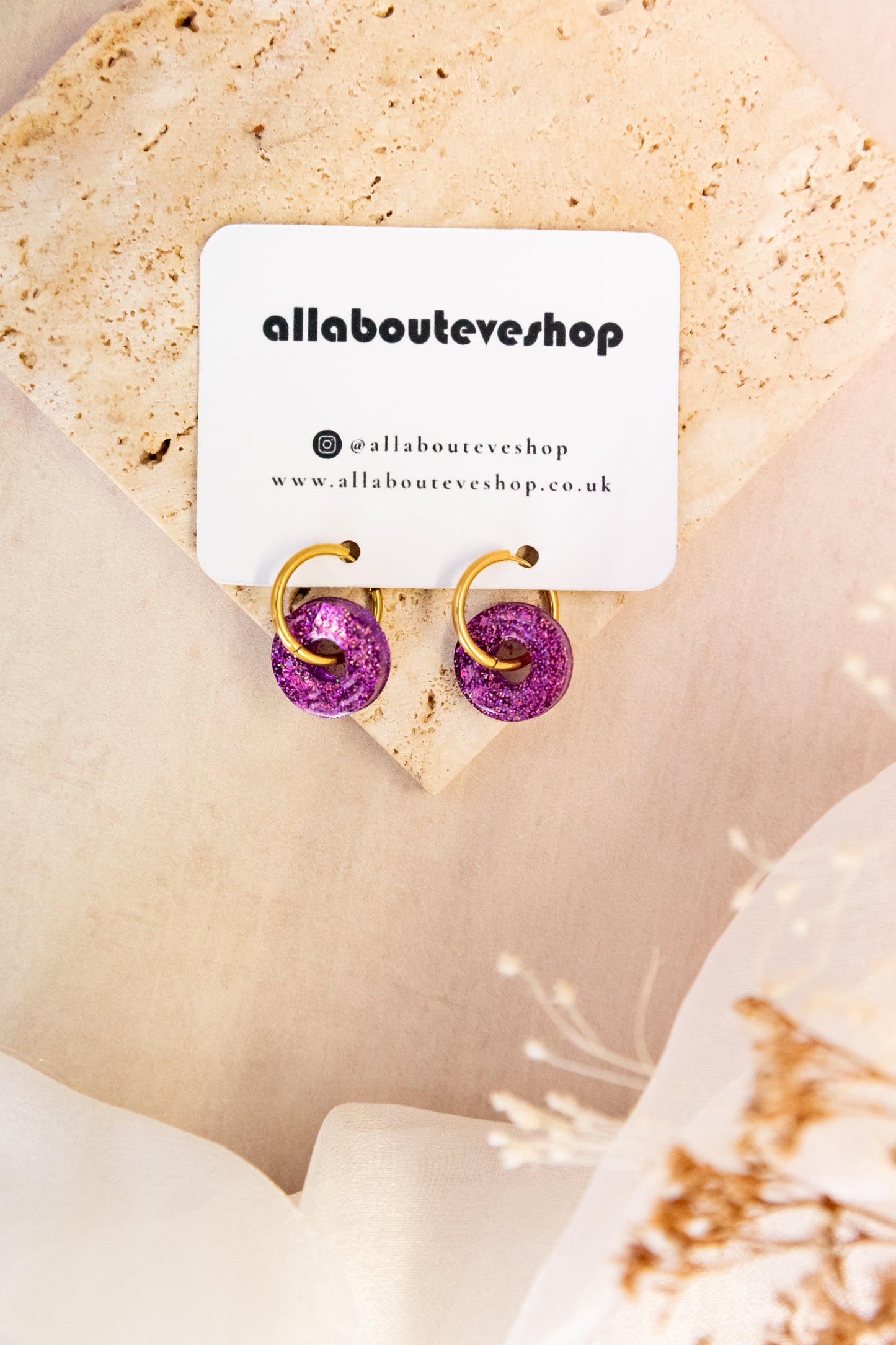 Gold and purple earrings on a beige stone with 'allabouteveshop' card.