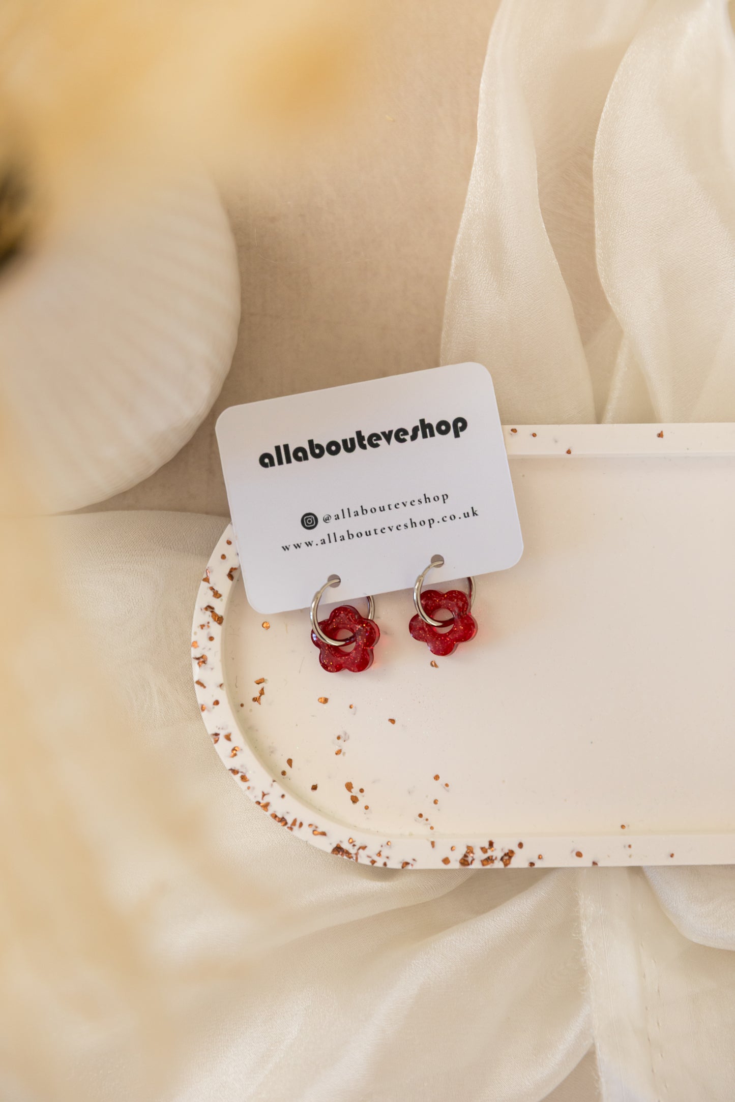 Red flower-shaped earrings on a white card with 'allabouteveryshop' branding, placed on beige fabric.