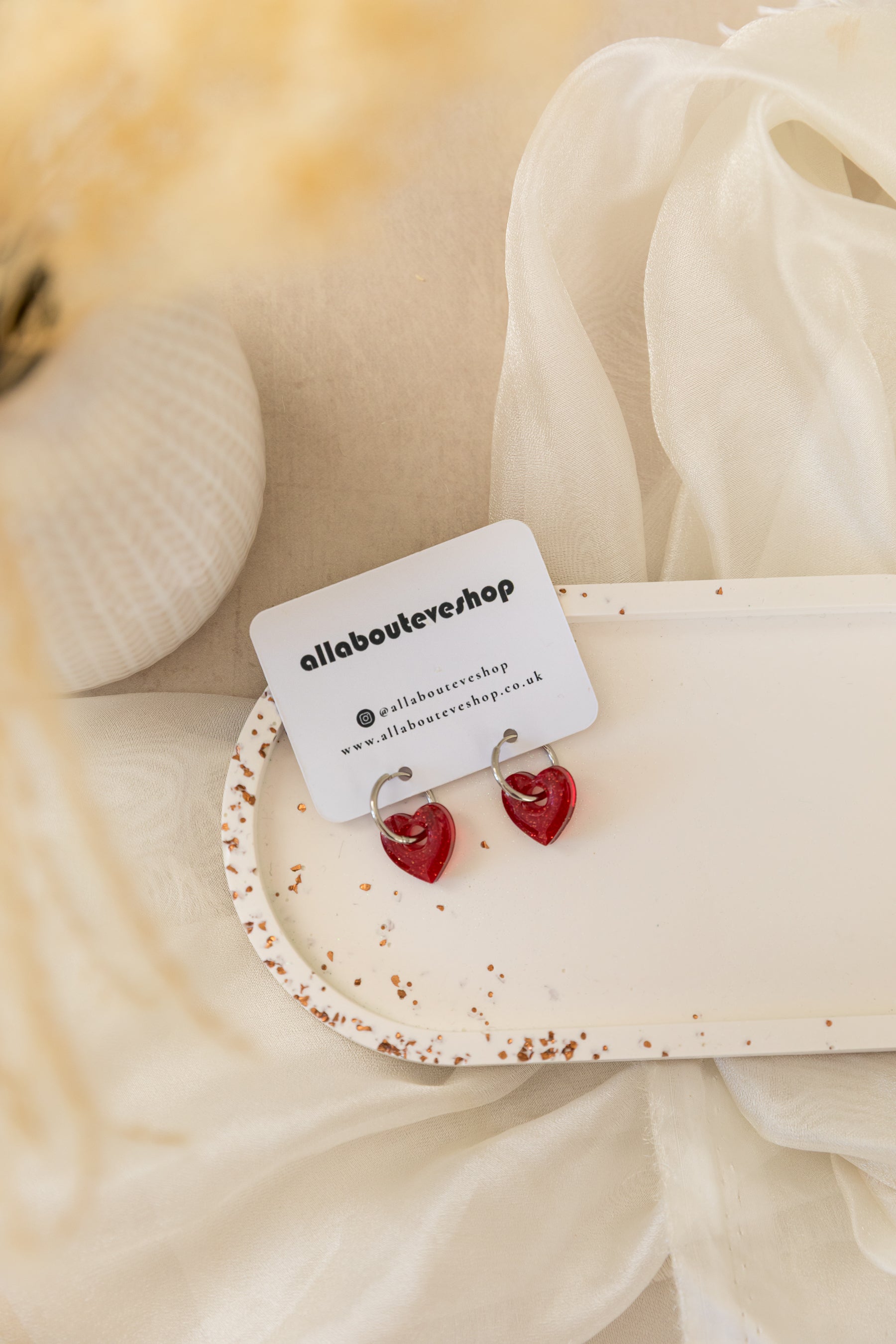 Heart-shaped earrings on a white card with 'allabouteve' branding, against a soft beige fabric background.