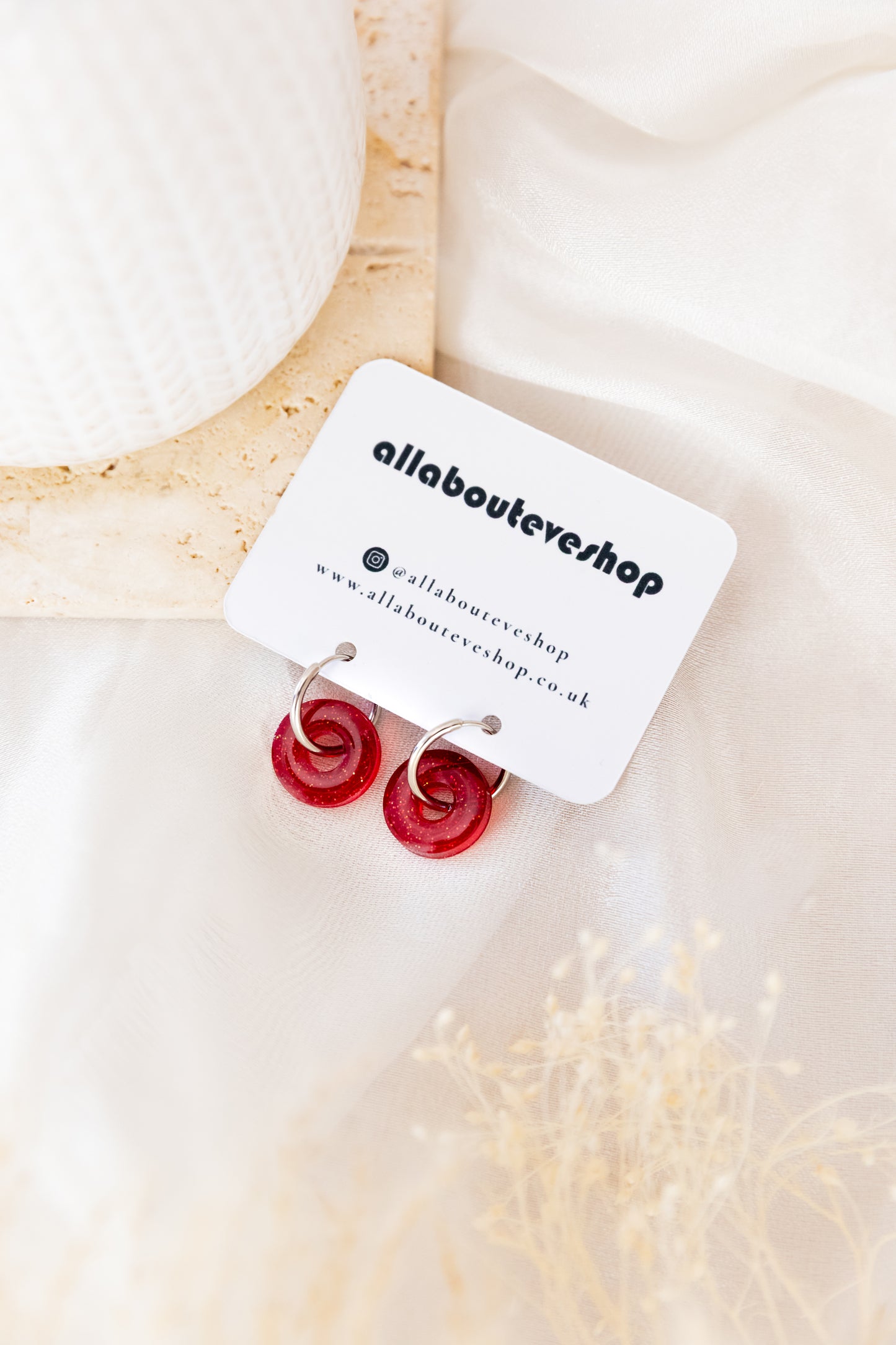 Red earrings on a white card with 'allabouteve' branding, placed on a soft beige fabric background.