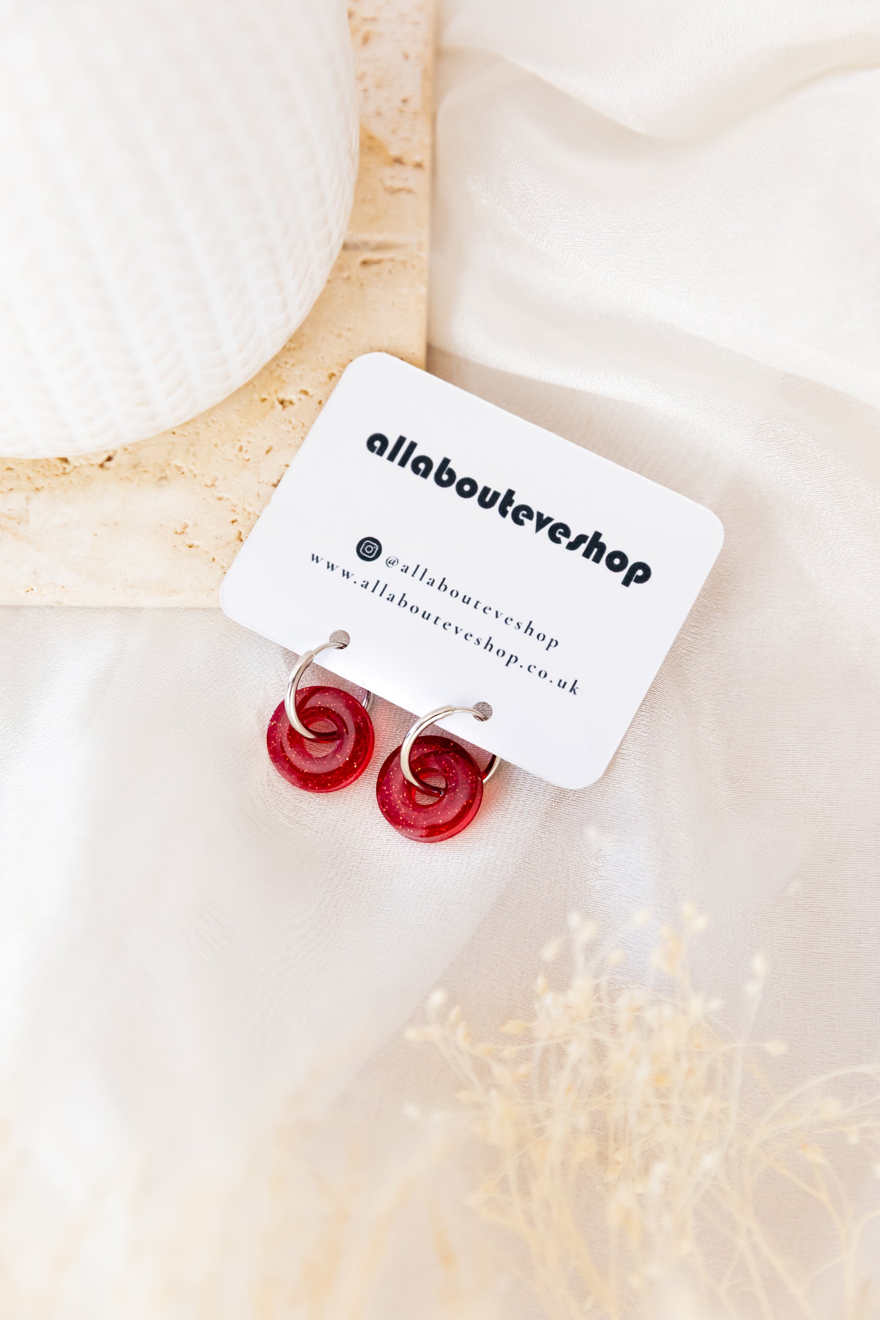 Red earrings on a white card with 'allabouteve' branding, placed on a soft beige fabric background.