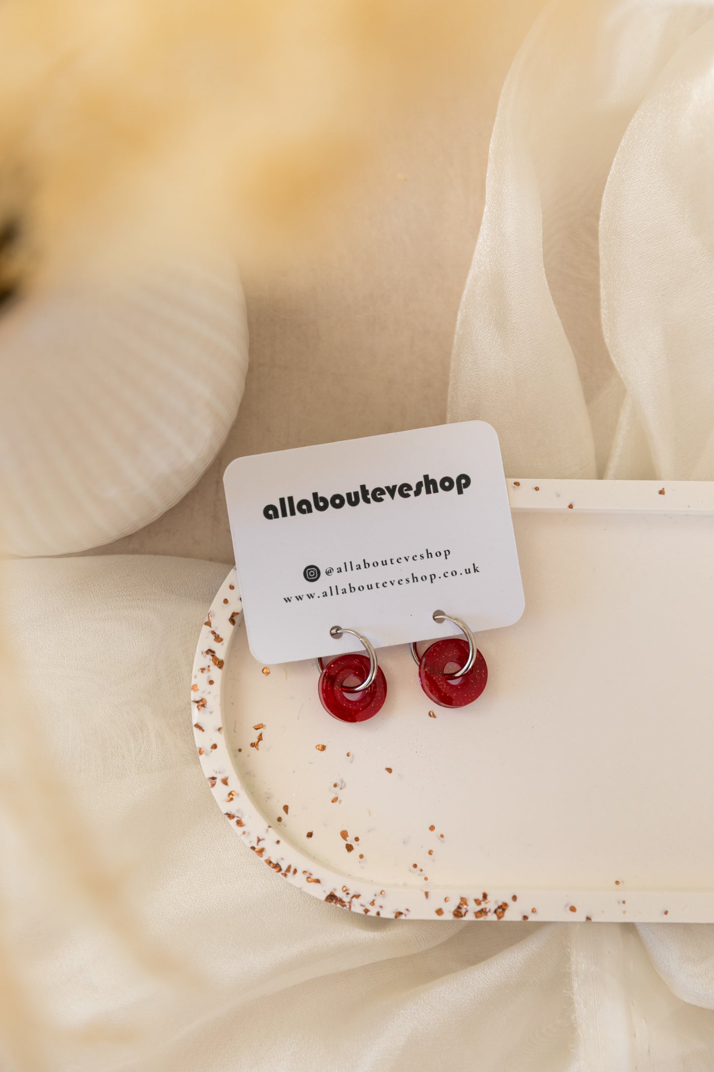 Red earrings on a white card with 'allaboutevershop' branding, placed on a beige fabric background.